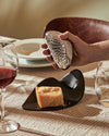 Metallic ergonomic grater by Alessi held in a hand over a wedge of parmesan cheese on a black plate.