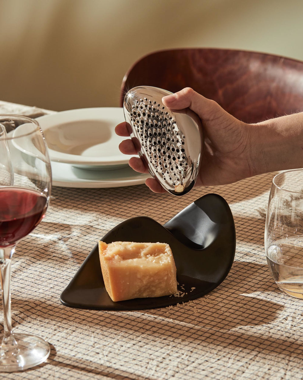Metallic ergonomic grater by Alessi held in a hand over a wedge of parmesan cheese on a black plate.