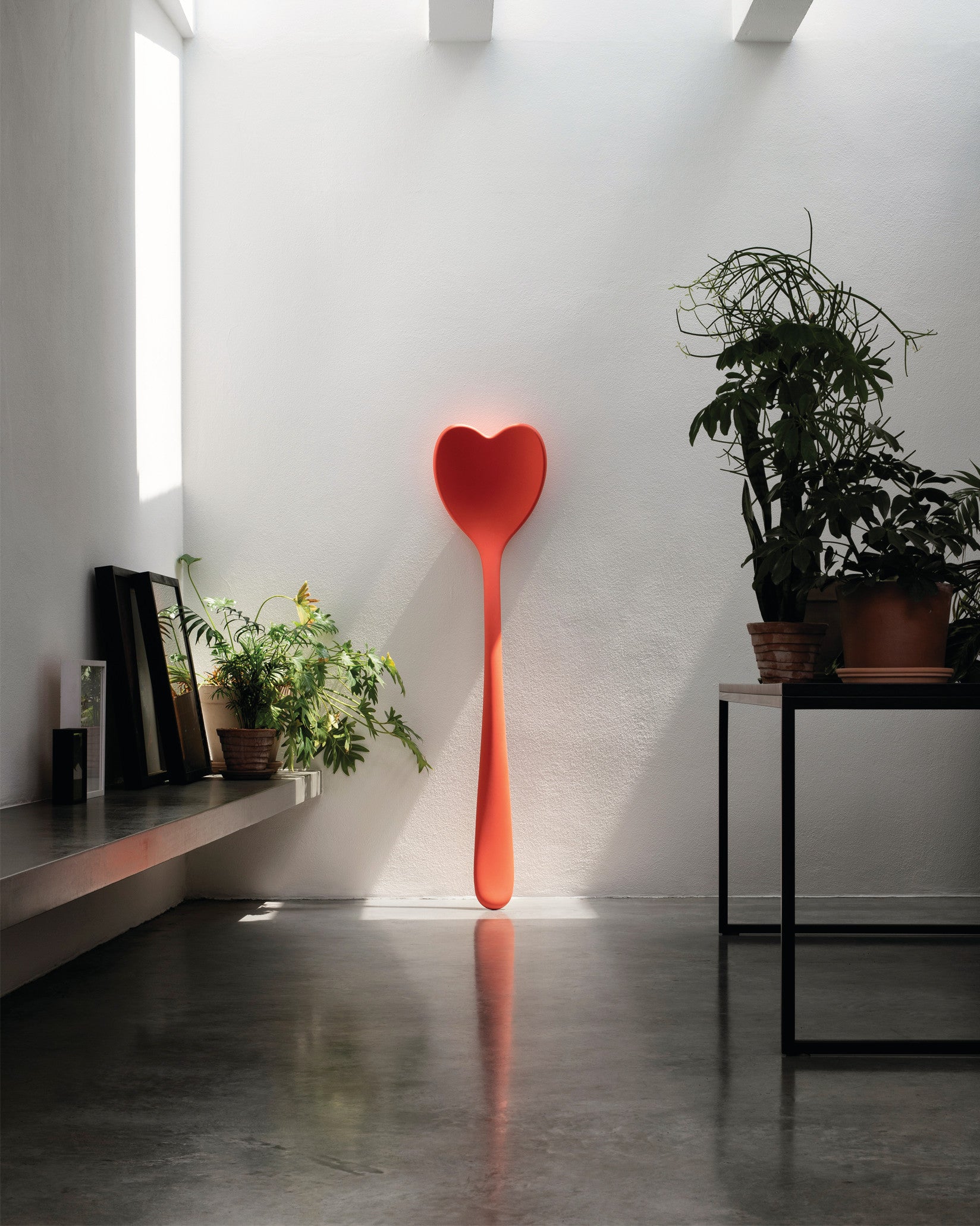 Large red heart-shaped decorative spoon by Alessi, standing upright against a white wall, reflecting on a concrete floor.