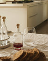 Wide clear drinking glass by Alessi, filled with water, on a patterned tablecloth with bottles and sliced bread, in a modern kitchen.