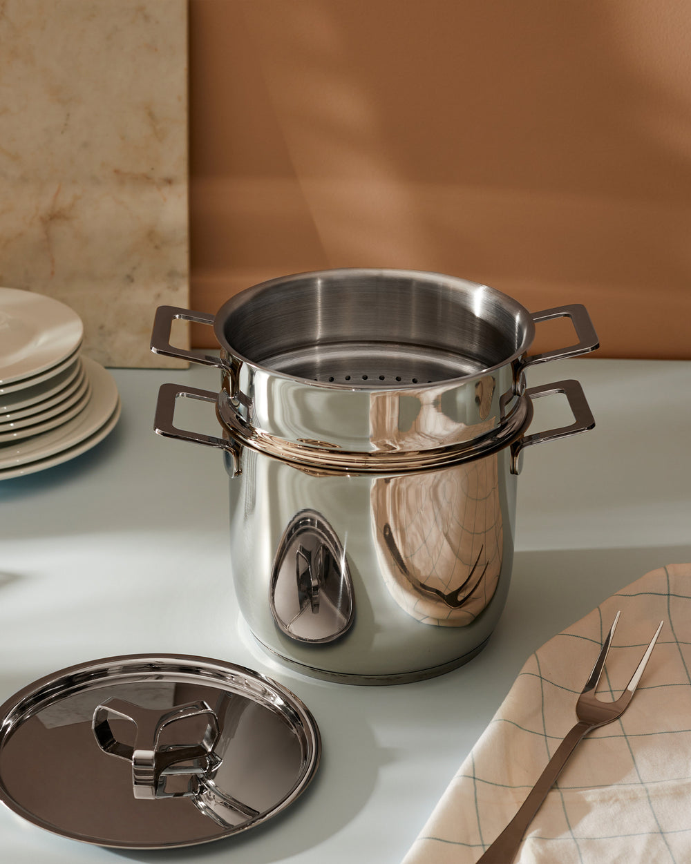 Stainless steel multi-tiered pot by Alessi with square handles, a perforated insert, and detached lid, on a light blue counter with a serving fork and stacked plates.