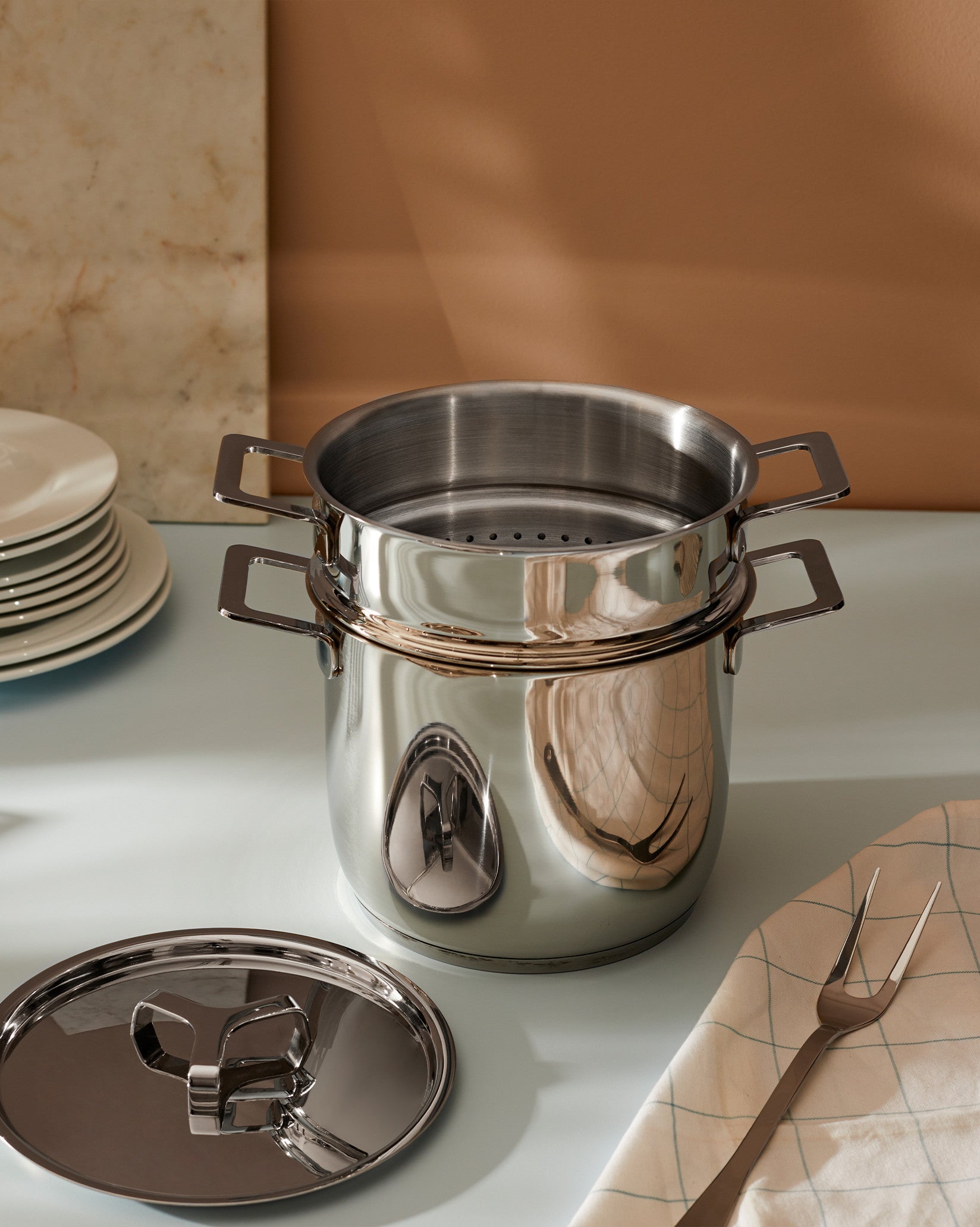 Stainless steel multi-tiered pot by Alessi with square handles, a perforated insert, and detached lid, on a light blue counter with a serving fork and stacked plates.