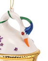 White ceramic swan hanging ornament by Alessi with a red jewel eye, orange beak, blue and green accents, purple flowers, and a golden textured base.
