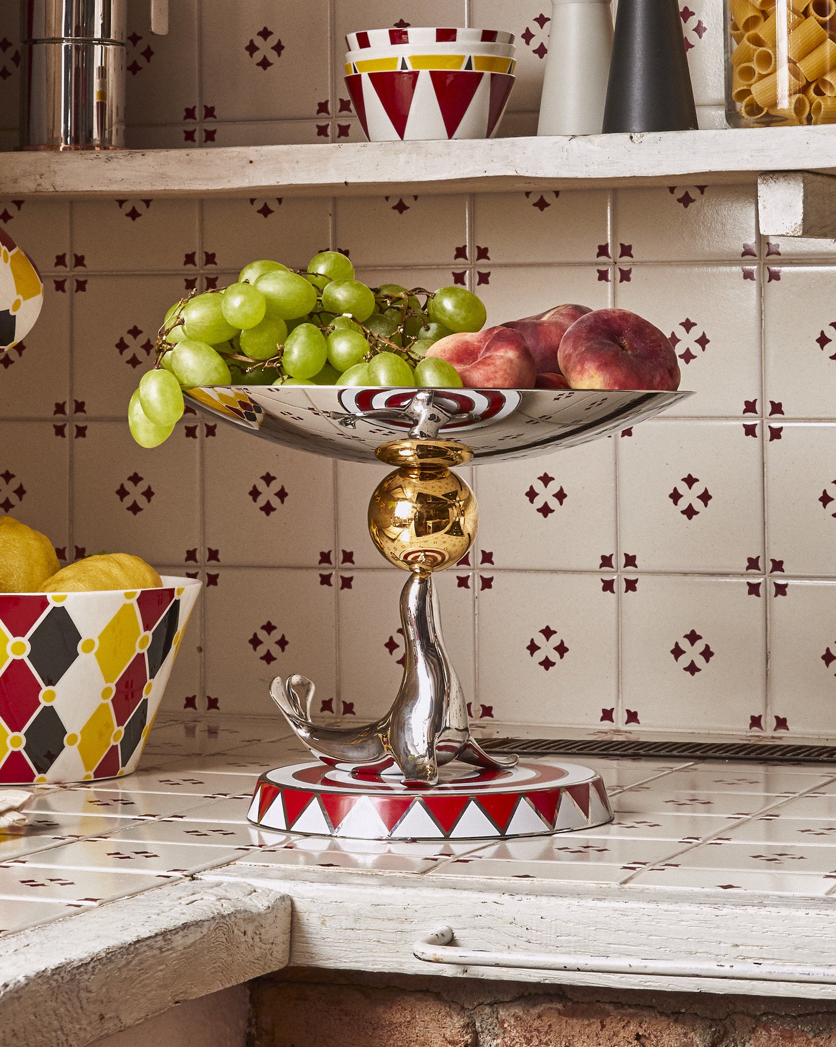 Silver-toned seal figure fruit bowl by Alessi, with a gold sphere and red and white patterned base.