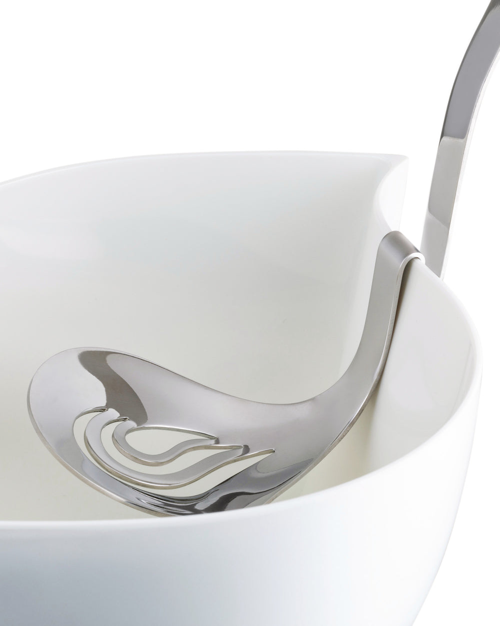 Silver slotted serving spoon by Alessi with wavy cutouts, elegantly resting in a white serving bowl.