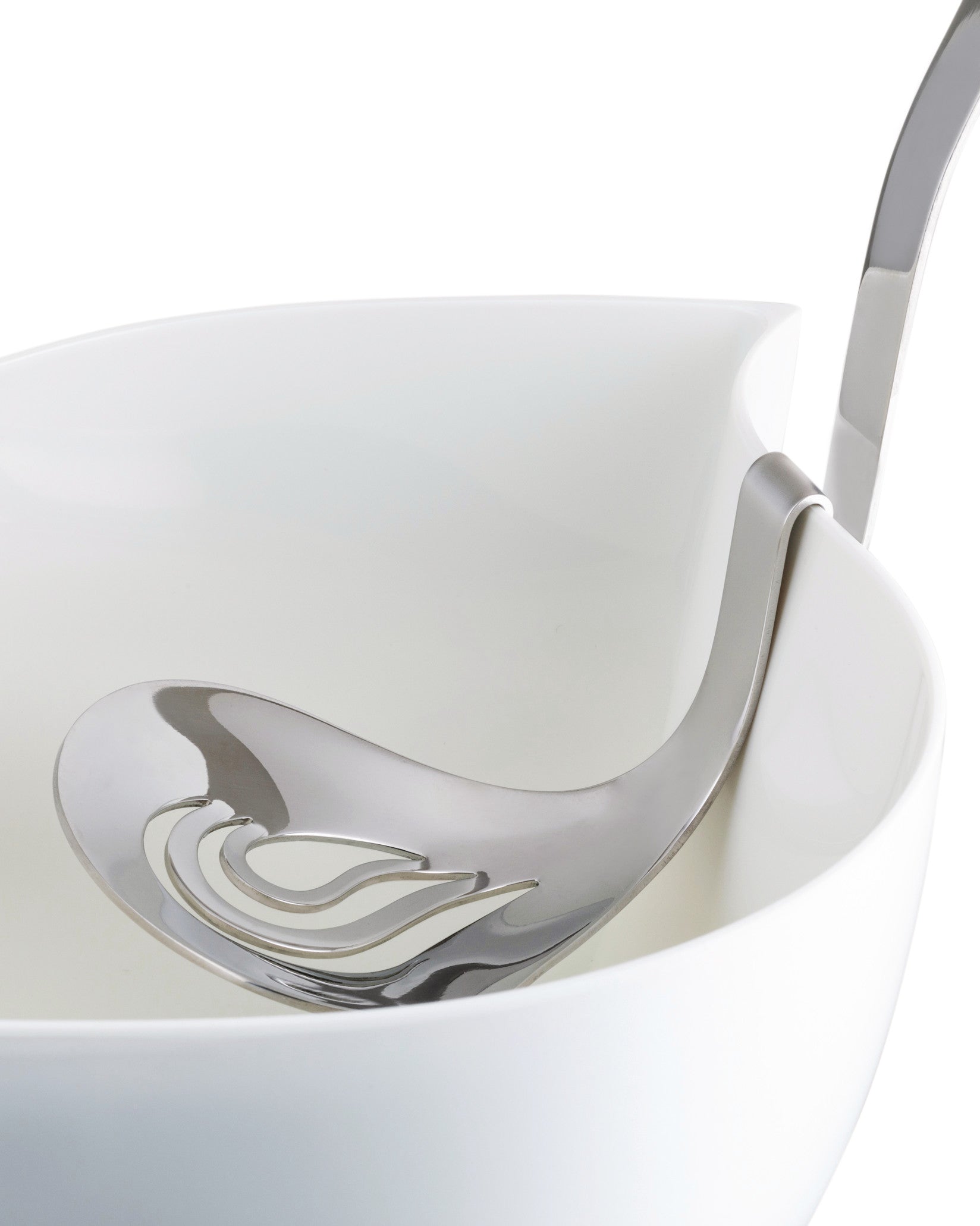 Silver slotted serving spoon by Alessi with wavy cutouts, elegantly resting in a white serving bowl.