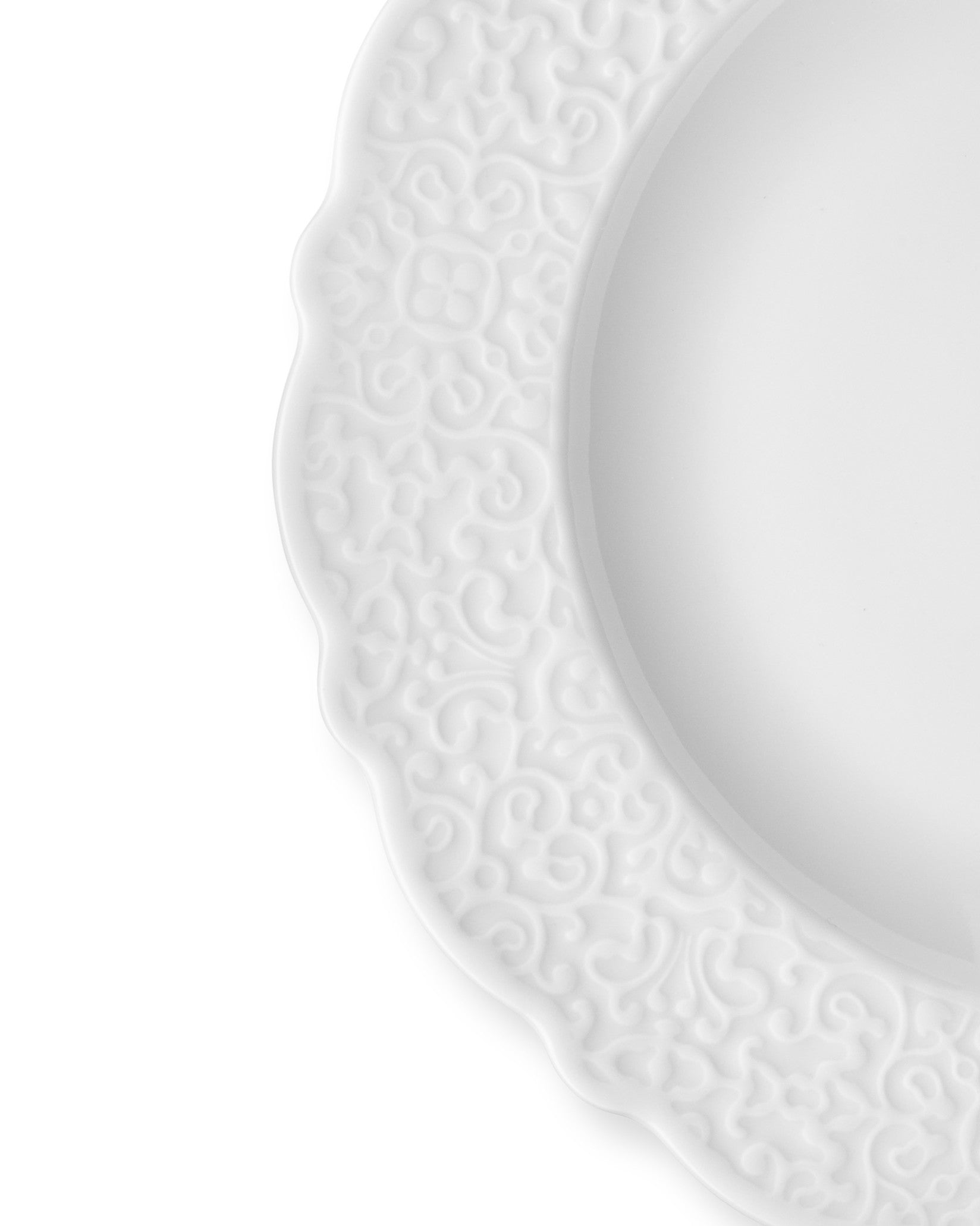 White ceramic plate by Alessi with a scalloped edge and embossed scrollwork pattern, partially visible.