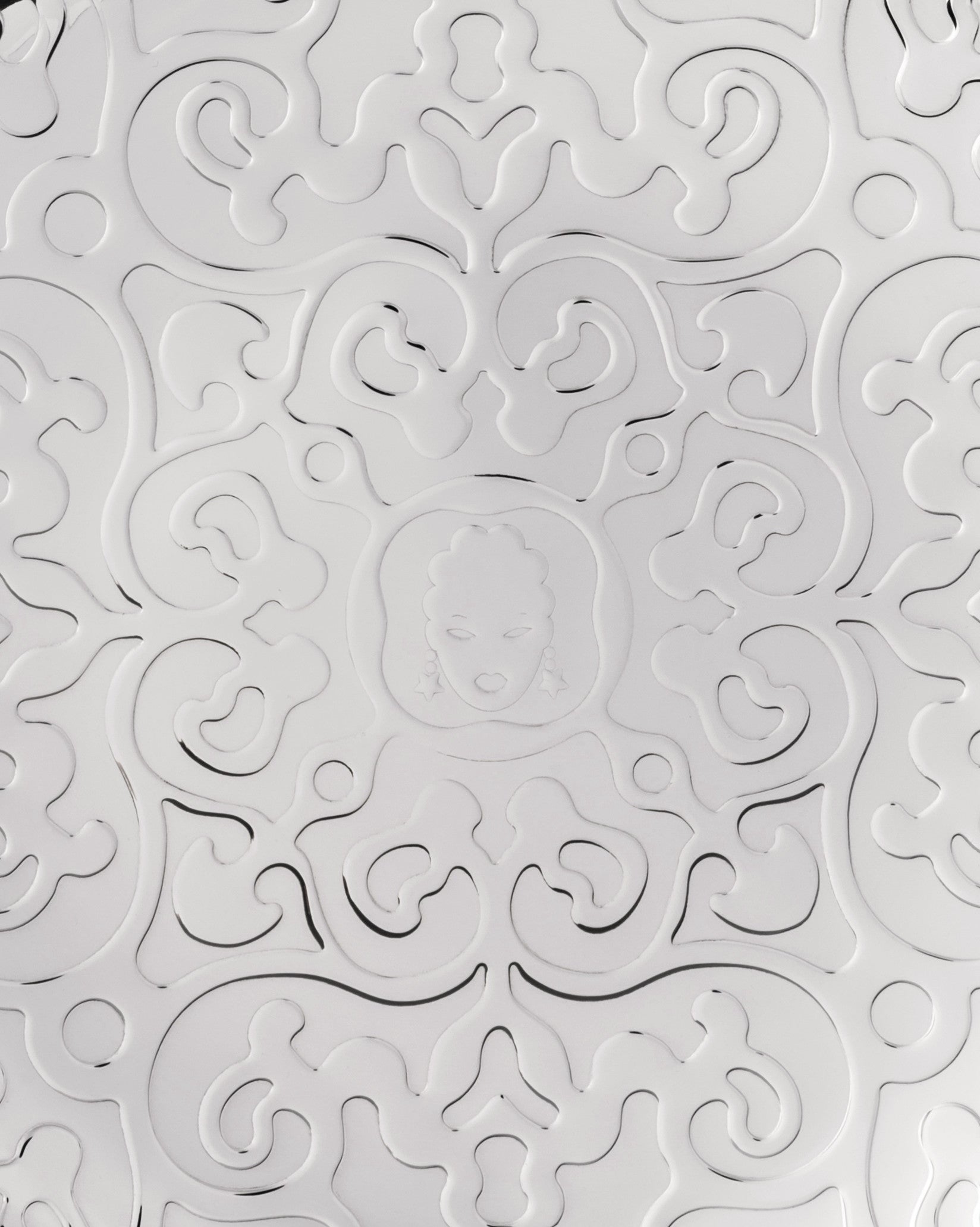 White embossed pattern by Alessi featuring swirling, floral-like designs and a central medallion with a stylized face wearing star earrings.