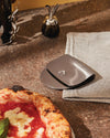 Polished stainless steel pizza cutter by Alessi, with an integrated cover, beside a fresh pizza and linen napkin.