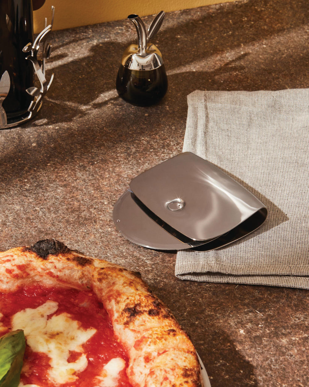Polished stainless steel pizza cutter by Alessi, with an integrated cover, beside a fresh pizza and linen napkin.