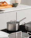 Stainless steel pots by Alessi with square loop handles and flat lids on a black induction cooktop in a modern kitchen.