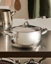 Polished stainless steel cooking pot by Alessi, with a lid and two handles on a metallic counter.