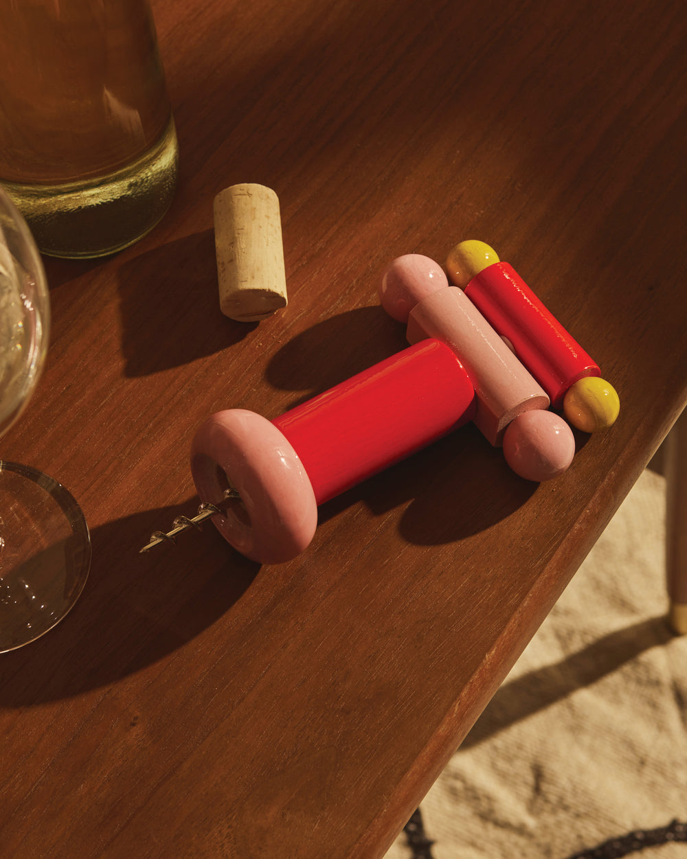 Red and pink ES17 corkscrew by Alessi, featuring yellow accents, on a wooden table with a wine bottle and cork.