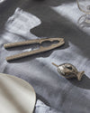 Polished silver fish-shaped condiment dish by Alessi, with textured scales, beside a matching nutcracker on grey linen.
