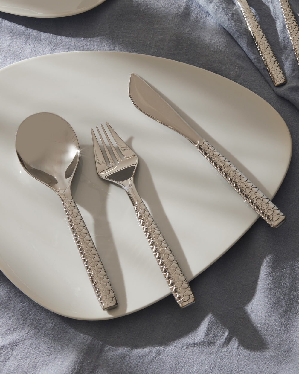 Silver-toned fish cutlery set by Alessi with textured scale-patterned handles on a white plate and blue linen tablecloth.