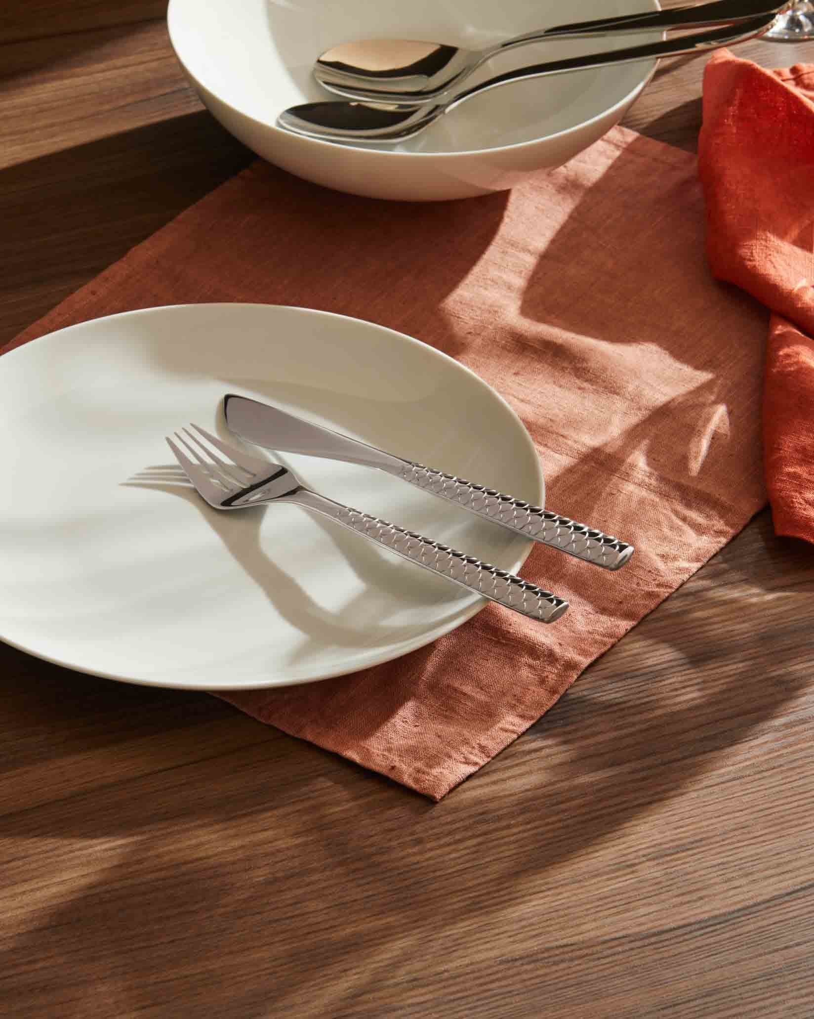 Silver-toned flatware by Alessi, featuring a fork and knife with geometric textured handles, on a white plate and orange placemat.