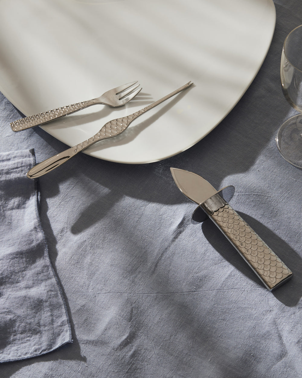 Silver-toned fish cutlery set by Alessi with a scale pattern, displayed on a white plate and blue linen.