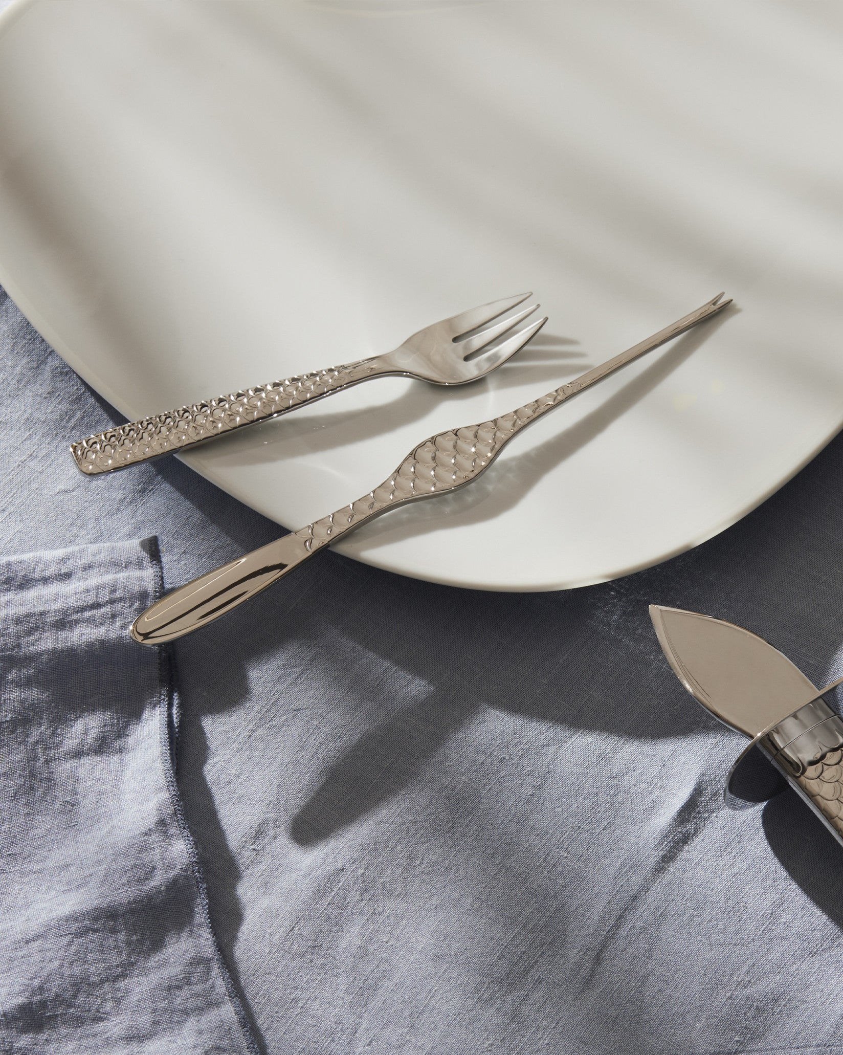 Silver Colombina fish cutlery set by Alessi featuring textured handles, placed on a white plate and blue linen.