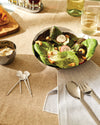 Stainless steel cutlery collection by Alessi, including a serving spoon, fork, and small decorative spoons, on a striped napkin.