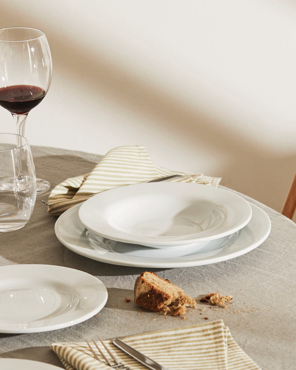 White ceramic dinnerware set by Alessi, styled on a grey linen table with striped napkins, bread, and wine.