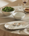 White ceramic plate and bowls by Alessi, with salad and serving utensils on a grey linen tablecloth.