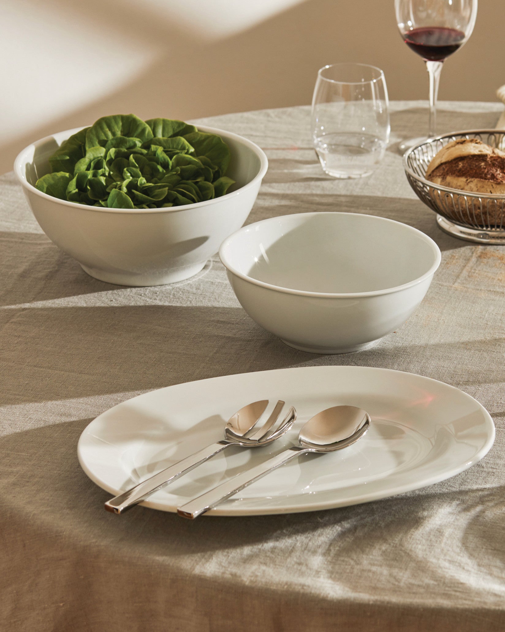 White ceramic plate and bowls by Alessi, with salad and serving utensils on a grey linen tablecloth.