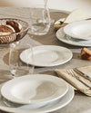 White plates and bowls by Alessi, set on a linen table with bread, glasses, and striped napkins.