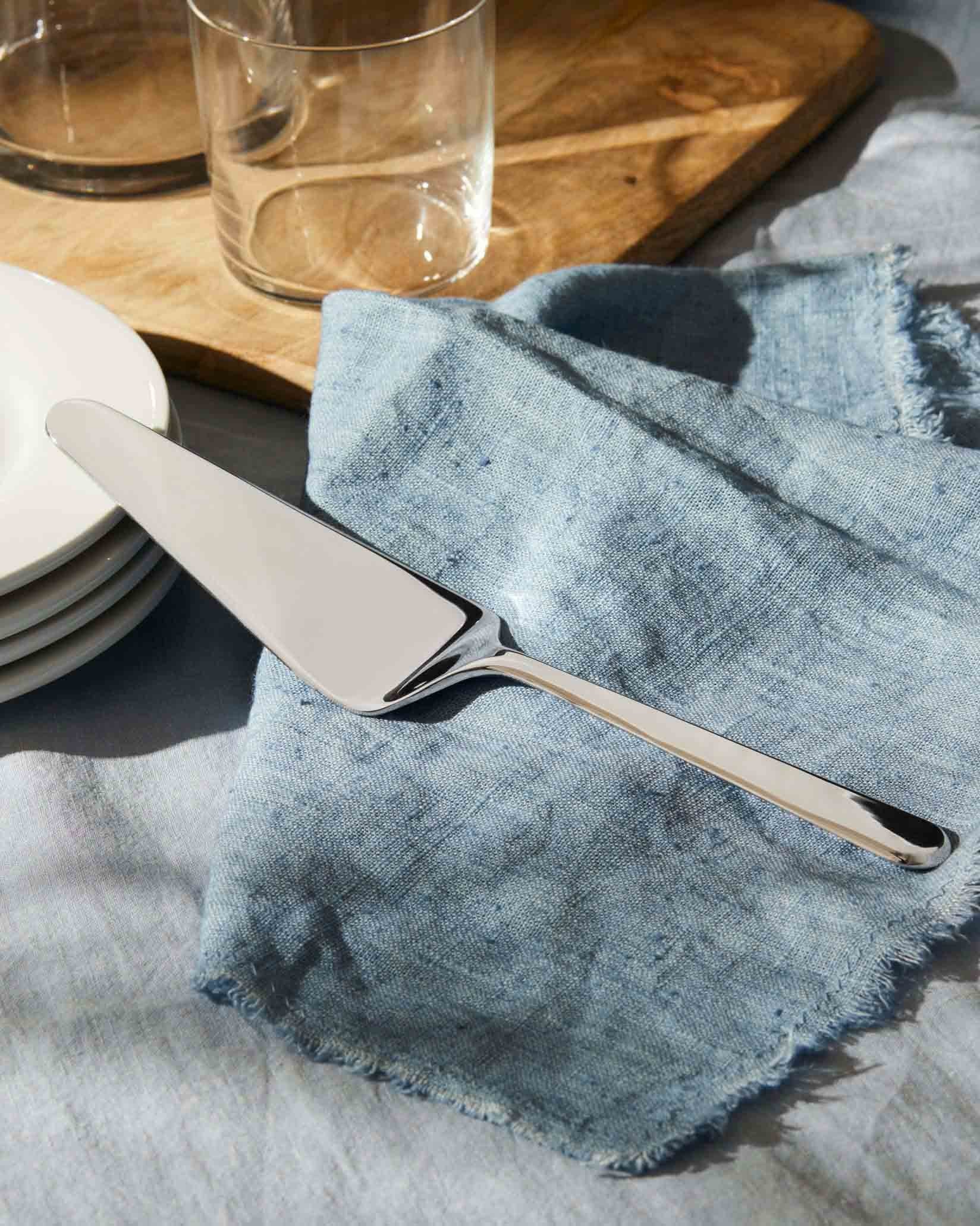 Silver-toned metal cake server by Alessi, with a sleek design, resting on a blue linen napkin next to white plates.
