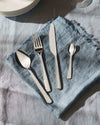 Silver stainless steel cutlery set by Alessi, neatly arranged on a blue linen napkin with frayed edges.