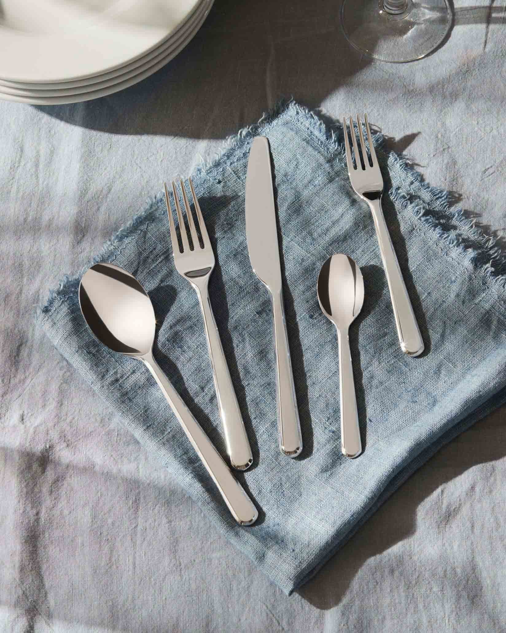 Polished silver five-piece cutlery set by Alessi on a textured blue linen napkin, with white plates.