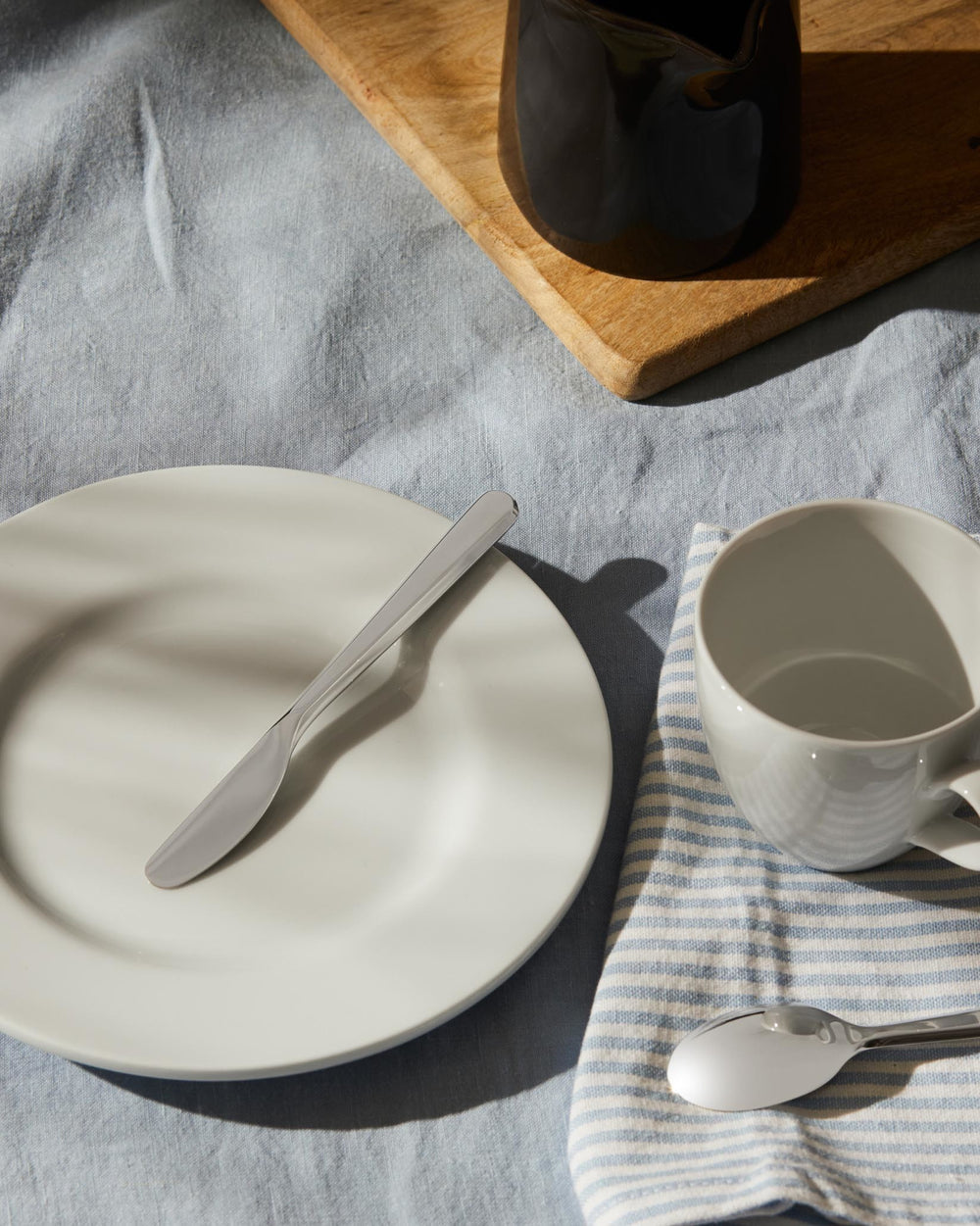 Sleek silver knife by Alessi resting on a white plate, with a blue and white striped napkin and white cup on a light blue tablecloth.