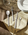 Modern silver flatware set by Alessi, featuring sleek, flat handles, arranged on a striped napkin with plates and glasses.