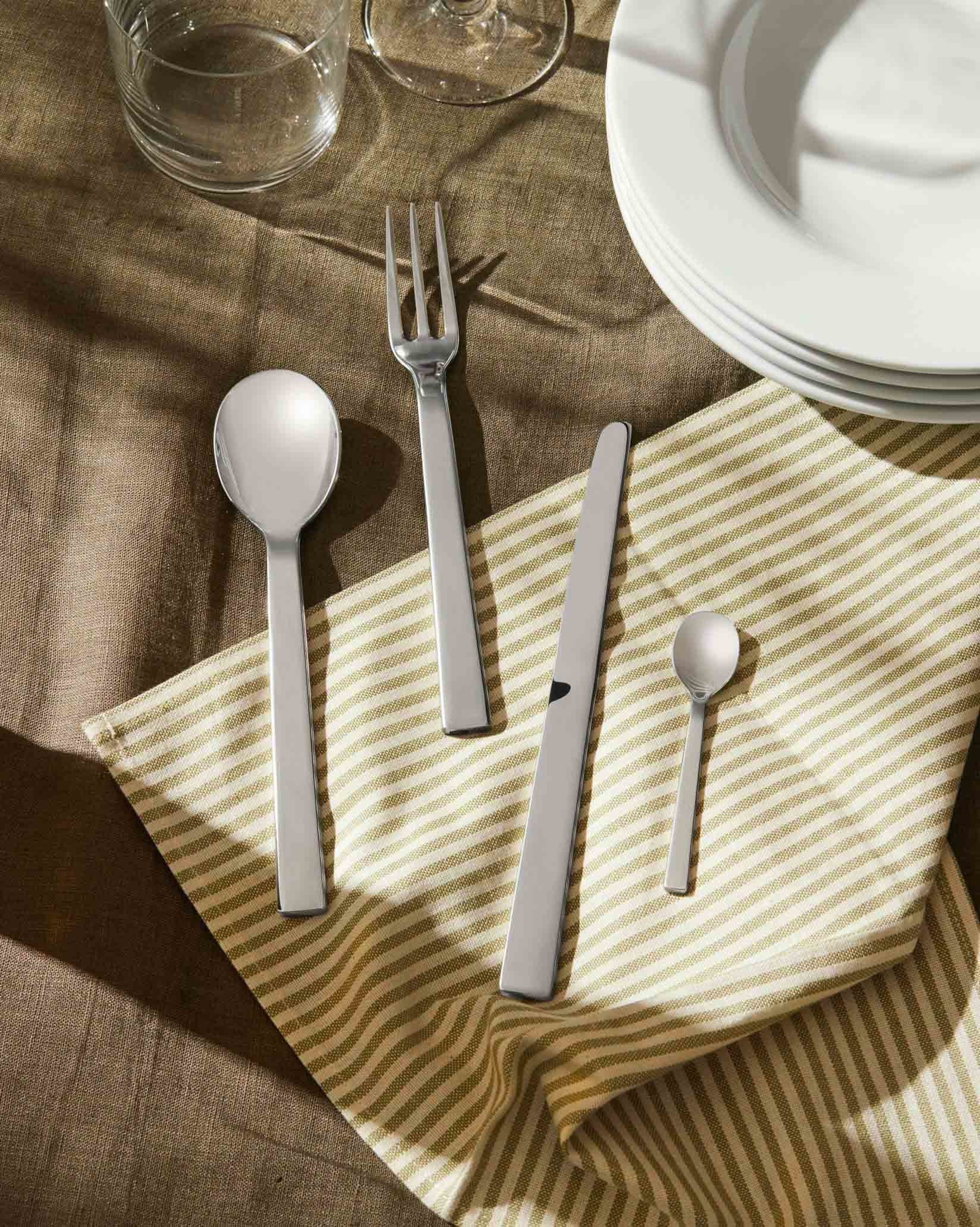 Modern silver flatware set by Alessi, featuring sleek, flat handles, arranged on a striped napkin with plates and glasses.