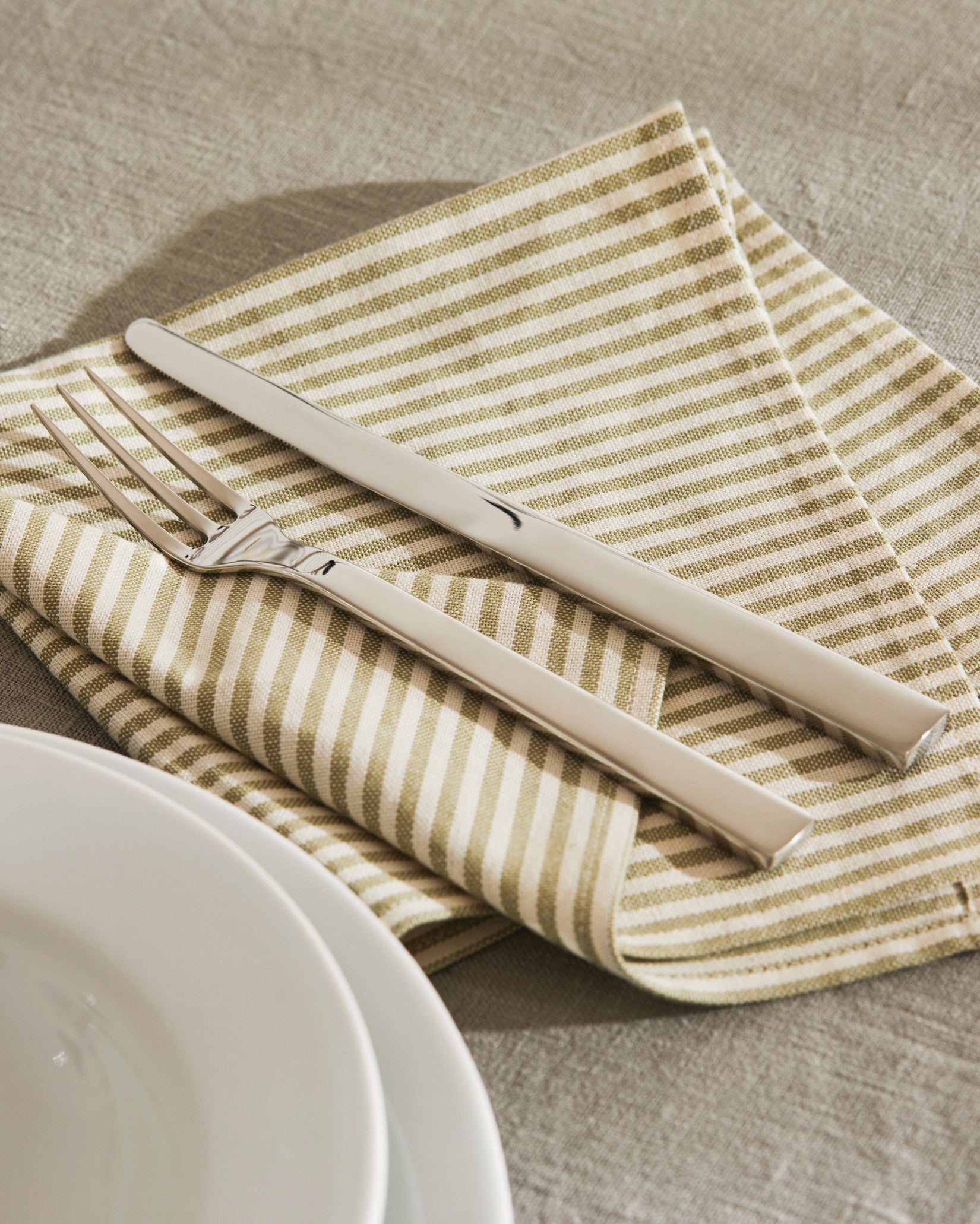 Sleek polished silver cutlery set by Alessi on a green and white striped napkin, with a white plate on a grey linen tablecloth.