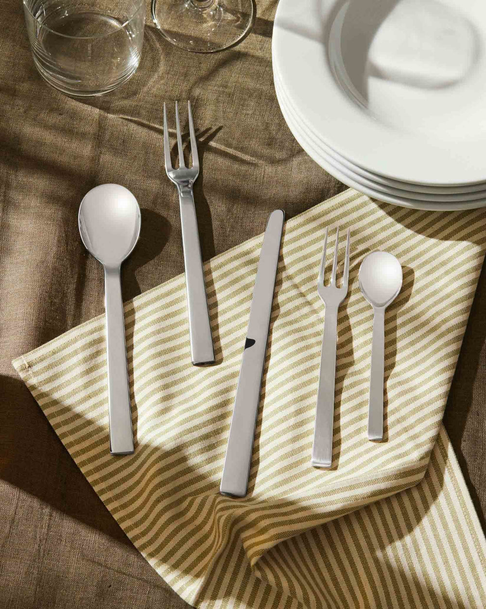 Silver flatware set by Alessi featuring five pieces with modern flat handles, on a striped napkin.
