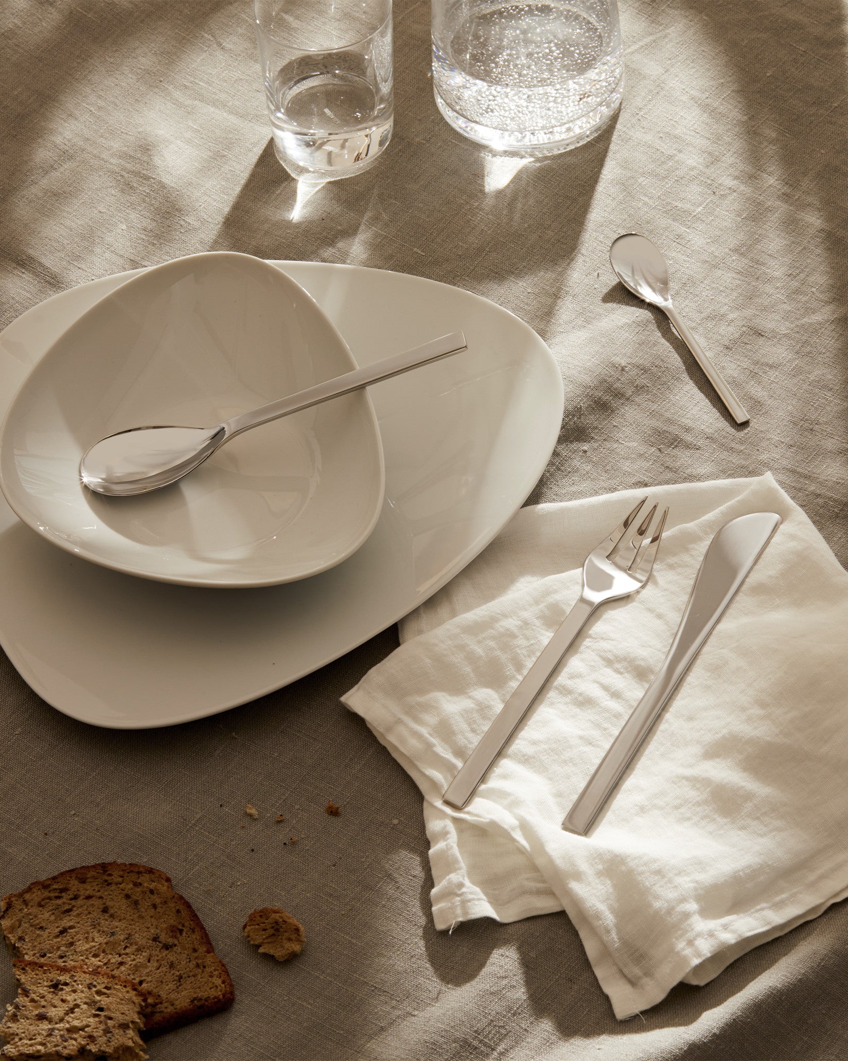 Colombina collection tableware by Alessi, including sleek stainless steel cutlery and white ceramic plates, on a beige linen tablecloth.