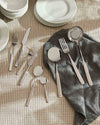 Polished stainless steel cutlery set by Alessi, including serving pieces, arranged on a grey linen napkin and plaid tablecloth.