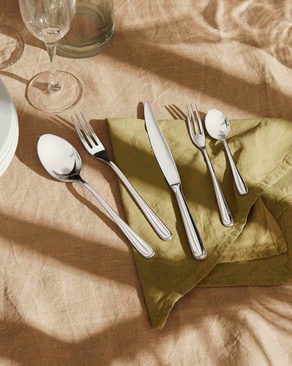Silver stainless steel cutlery set by Alessi, including a knife, two forks, and two spoons, on a green linen napkin.