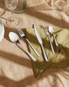 Silver cutlery set by Alessi, neatly arranged on a green napkin on a brown linen tablecloth.