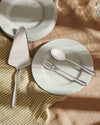 Silver metallic Caccia cutlery by Alessi, featuring a cake server, fork, and spoon on a light green plate with a striped napkin.