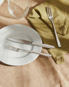 Sleek silver-toned cutlery set by Alessi on a white plate, green napkin, and linen tablecloth.