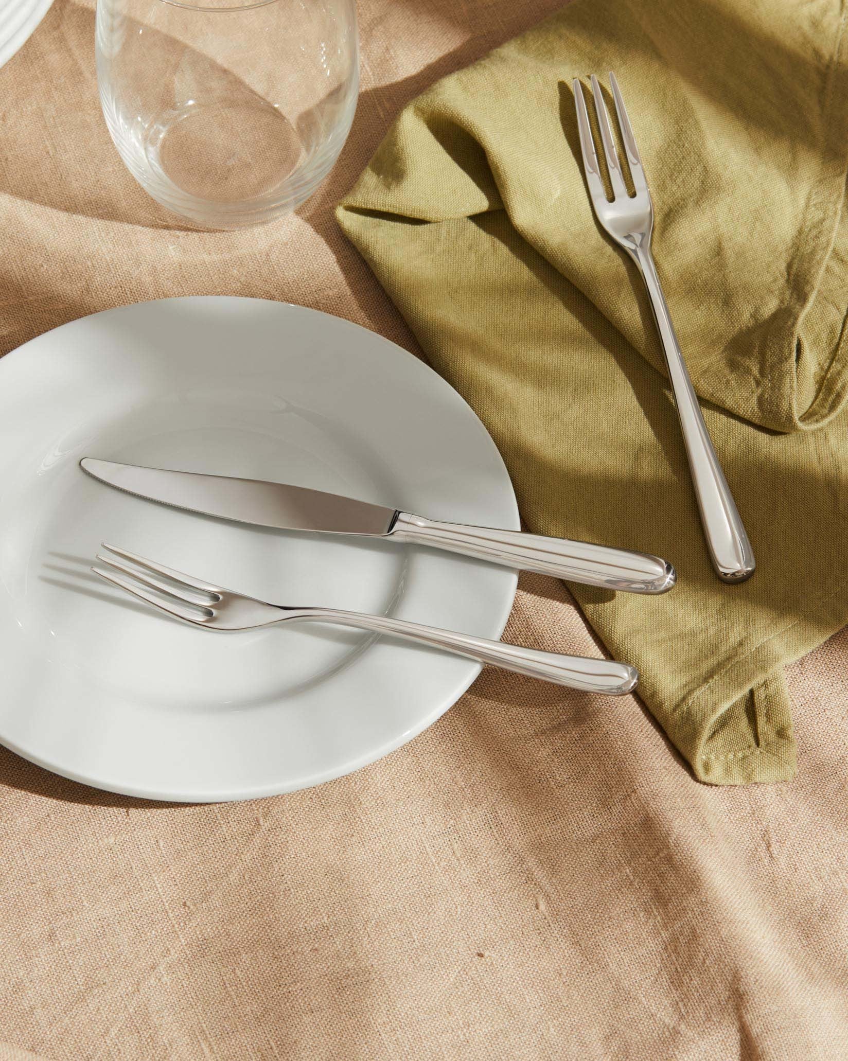 Caccia stainless steel cutlery by Alessi, with a knife and fork on a white plate and green linen napkin.
