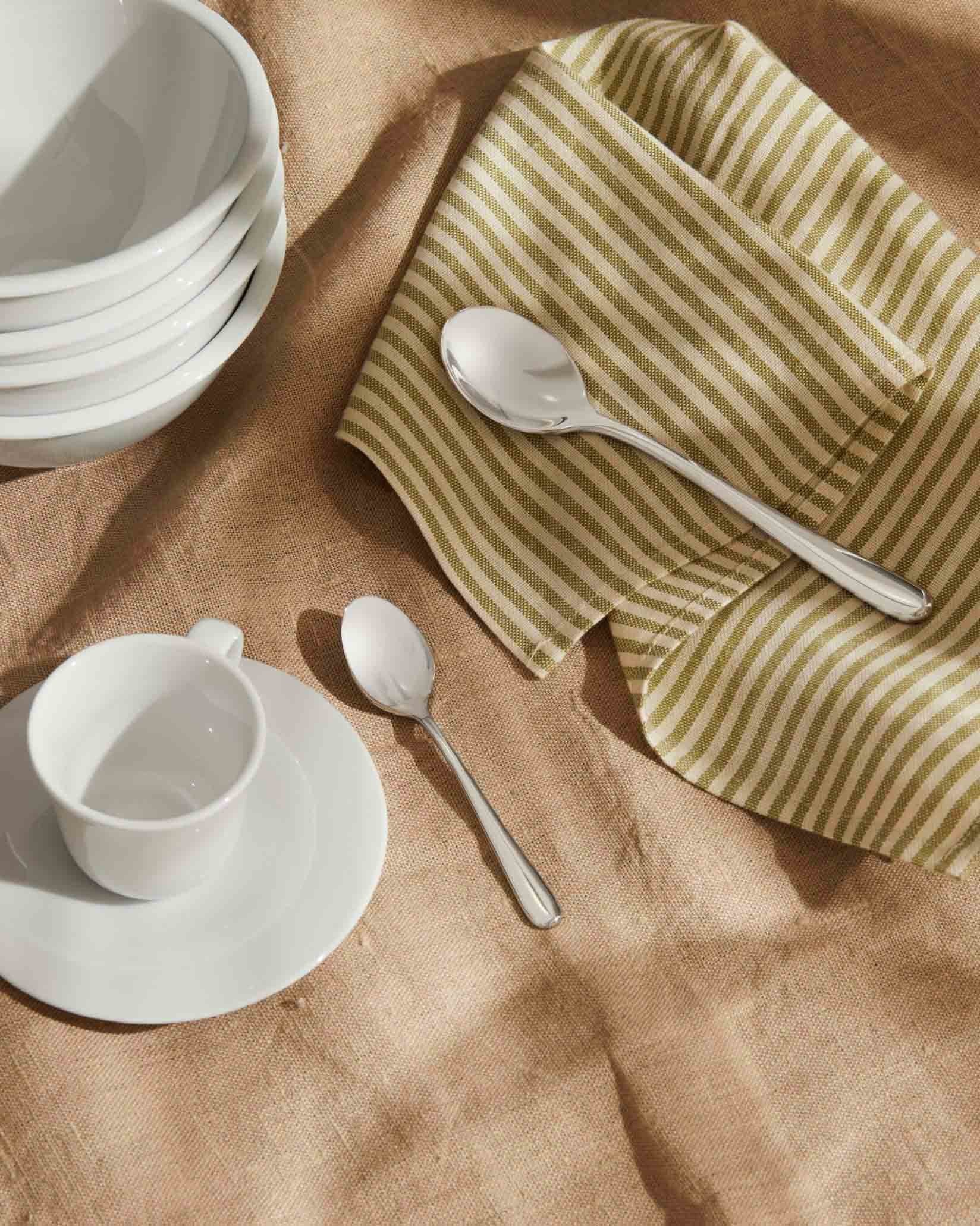 Two silver-toned spoons by Alessi, resting on a striped napkin and natural linen tablecloth with white dishes.