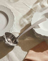 Polished silver serving ladle by Alessi, angled on a beige linen tablecloth with stacked white scalloped plates.