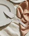 Silver cake server by Alessi with ornate embossed scrollwork handle, resting on a decorative white plate.