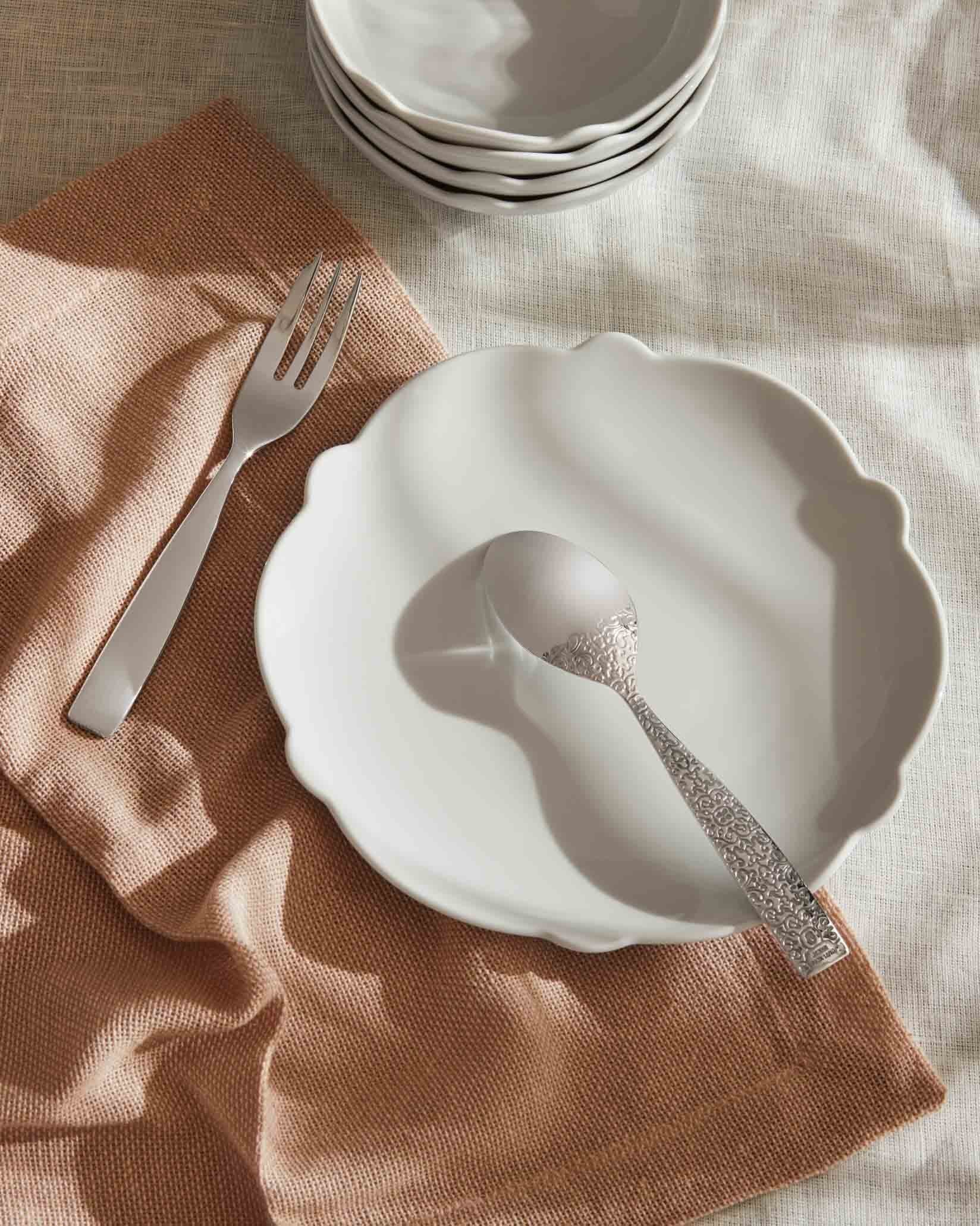 Scalloped white dinner plate by Alessi, with a patterned silver spoon and plain fork on a brown napkin.