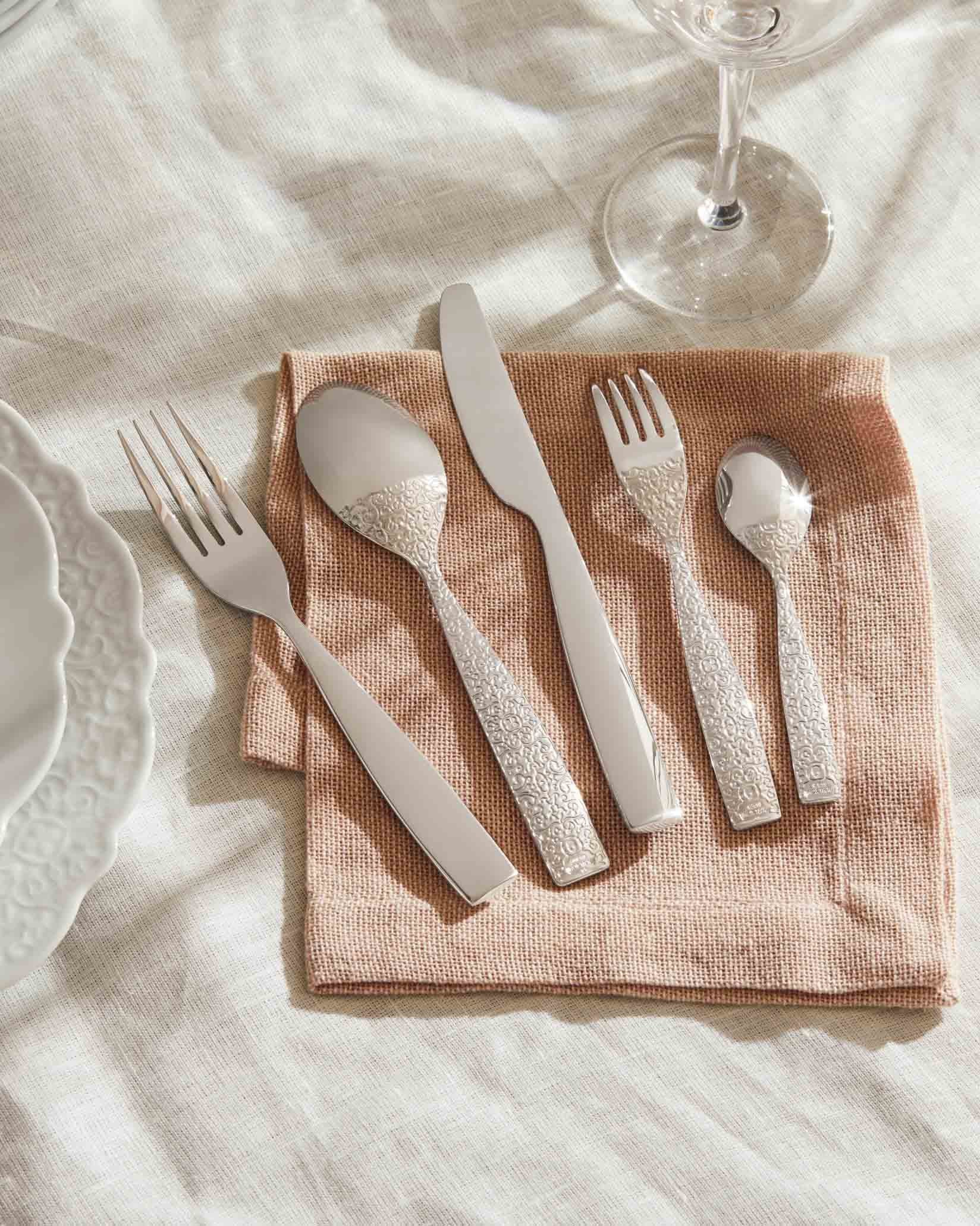 Silver cutlery set with ornate handles by Alessi, placed on a peach napkin and linen cloth.