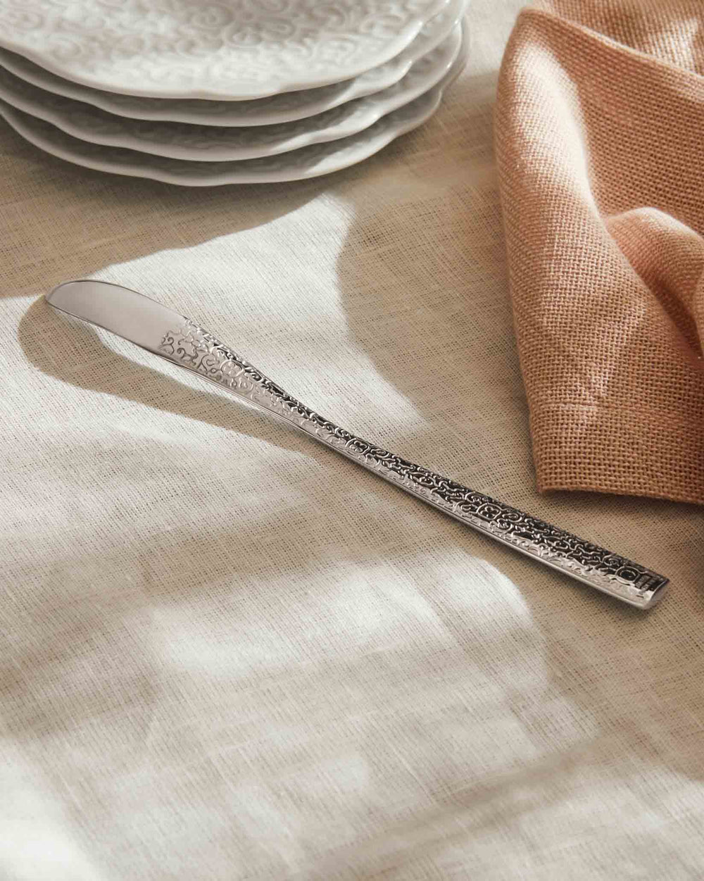 Silver Dressed butter knife by Alessi, with an intricately patterned handle, resting on a textured linen tablecloth.