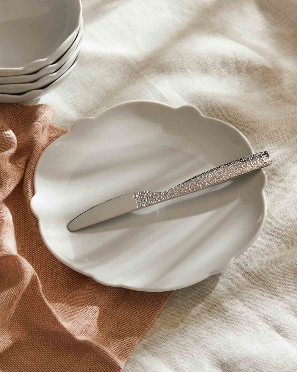 Scalloped white plate by Alessi, with an ornate silver knife on a cream linen cloth and rust napkin.