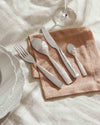 Stainless steel cutlery set by Alessi with ornate patterned spoon handles and smooth knife and fork handles, arranged on a rust-colored linen napkin.